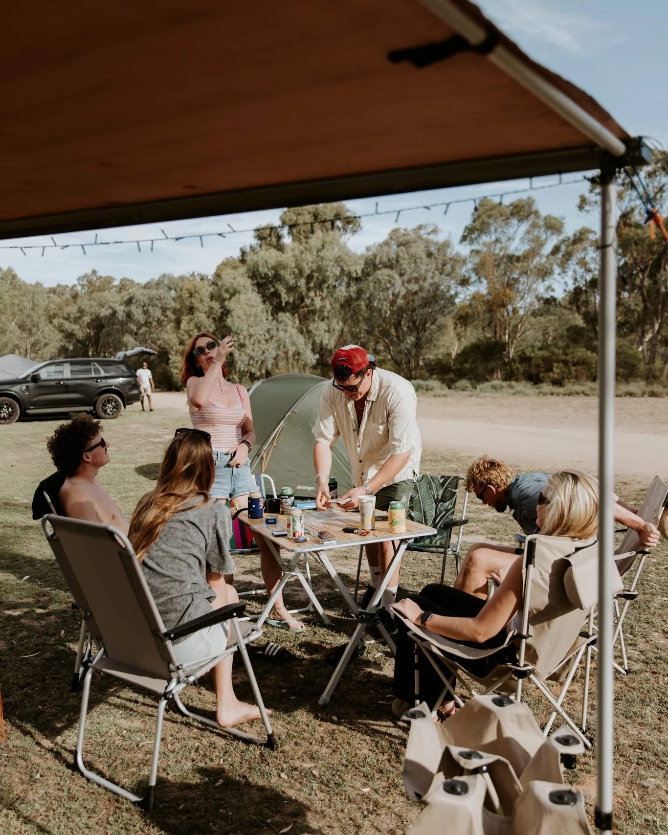 Friends gathered around a camping table under an awning, with a tent behind them