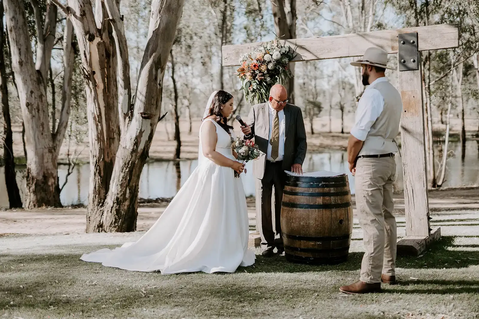 A wedding ceremony at the red gum arbour beside the lagoon, with a wine barrel between the couple